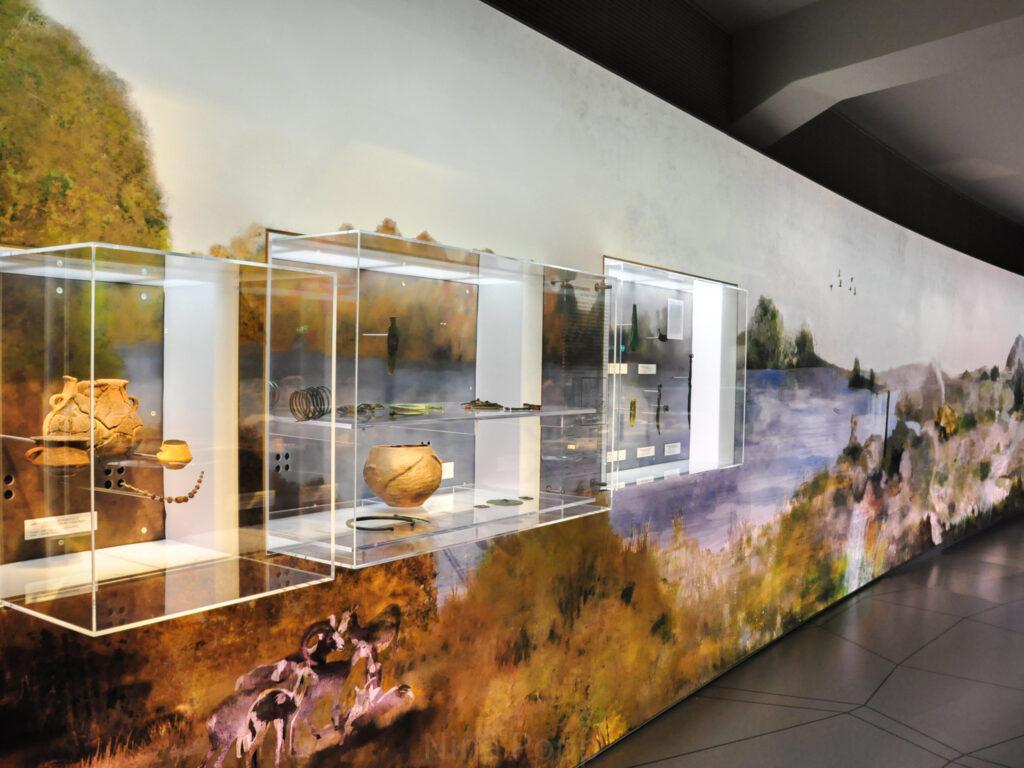 Glaskästen mit Keramikgefäßen an einer illustrierten, von hinten beleuchteten Wand.