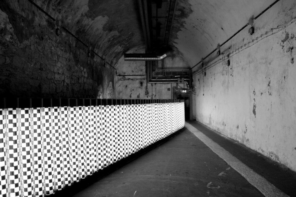 ECHO/Turning the light around, von VROEGOP/SCHOONVELD im Lichtkunstmuseum Unna (2017). Rotierende Leuchtröhren erstrecken sich von vorne links nach hinten rechts und ziehen ein langes Band hinweg, das im schwarz-weiß Schachbrettmuster ist.