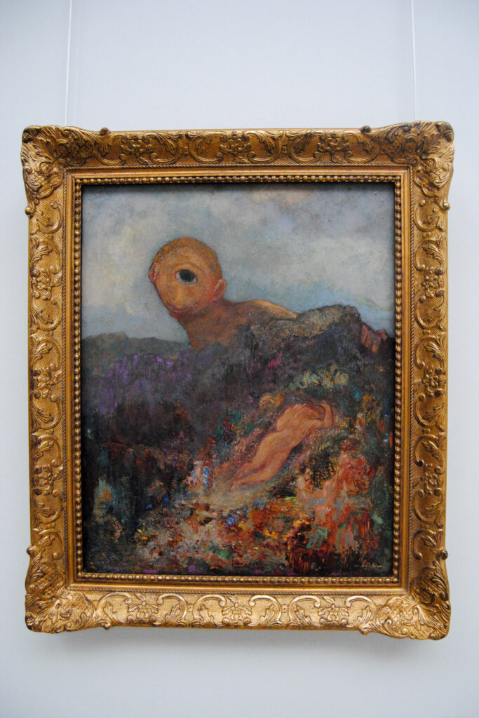 Odilon Redons Werk Le cyclope veranschaulicht im symbolistischen Stil: bunt und überlappende, verwaschene Pinselstriche, ein Zyklop, der Hals aufwärts über einen Berg hinauslugt. Auf dem Berg liegt eine Frau.